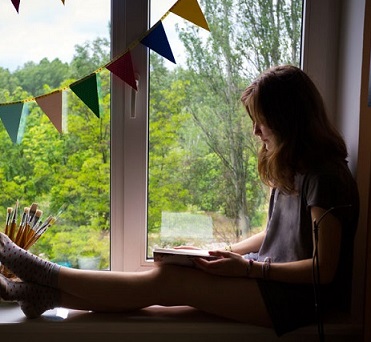 Una niña lee en su habitación frente a la ventana.